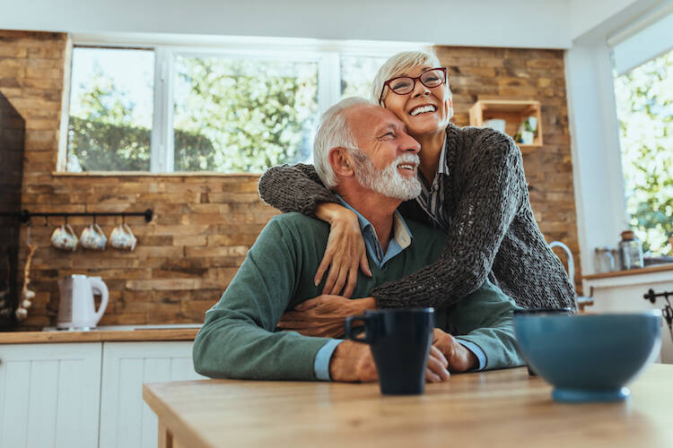 Retired elderly couple sitting at kitchen table happily hugging each other