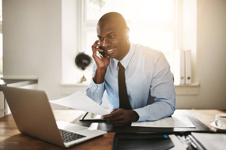Fiduciary financial investment advisor sitting at a desk, looking at a laptop and making a phone call on cellphone